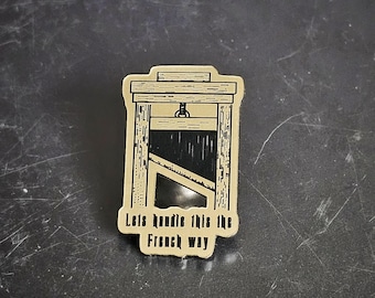 May include: A tan and black guillotine-shaped pin with the text "Lets handle this the French way". The pin features a detailed illustration of a guillotine, a historical execution device.