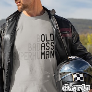 May include: A light gray t-shirt with the words "BOLD BADASS SUPERHUMAN" in a stacked design. The image also shows a black motorcycle helmet with a checkered pattern and the text "Charly Artly". The person is wearing a black leather jacket.