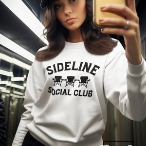 May include: White sweatshirt with black text that reads "Sideline Social Club" and a graphic of three black director's chairs. The sweatshirt is being worn by a person wearing a black baseball cap.