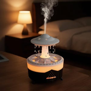 May include: A white and black UFO-shaped humidifier with a central pillar emitting a vertical stream of mist. The device features a saucer-like top, a glowing orange light band, and a base with control buttons, creating a rain-like water effect.