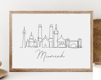 May include: A black and white line drawing of the Munich skyline, including the Frauenkirche, the Neues Rathaus, and the Olympic Tower. The drawing is framed in a light wood frame and is hanging on a white wall.