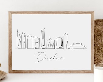 May include: A black and white line art print of the Durban skyline with the word "Durban" written below. The print is framed in a light brown wood frame and is displayed on a white surface.