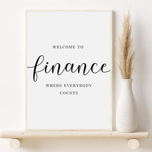 May include: A white framed print with black text that reads "Welcome to finance where everybody counts". The print is sitting on a light wood shelf with a vase of dried pampas grass.
