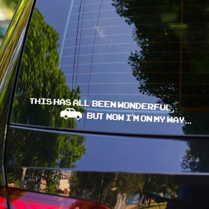 Op de afbeelding: Een witte vinyl auto sticker op de achterruit van een zwarte auto. De sticker zegt: "THIS HAS ALL BEEN WONDERFUL, BUT NOW I'M ON MY WAY..." met een kleine auto afbeelding. De auto staat buiten geparkeerd.