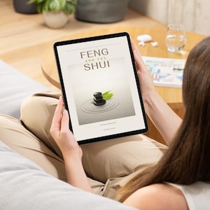 Könnte beinhalten: Eine Person hält ein schwarzes Tablet, auf dem das Cover eines Buches mit dem Titel "FENG AND YOU SHUI" angezeigt wird. Das Cover zeigt ein Zen-Garten-Design mit gestapelten schwarzen Steinen und grünen Blättern. Die Person trägt ein weißes Oberteil und eine beige Hose.