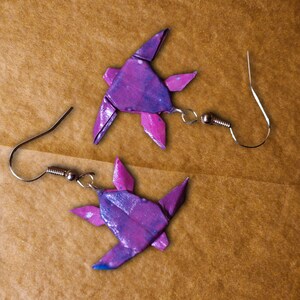 May include: A pair of origami turtle earrings with a purple and blue marbled design. The earrings are made of paper and have silver hooks.