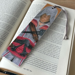 May include: A bookmark featuring a detailed illustration of a character in a hooded cloak, with red and gray accents. The bookmark is placed in an open book, with a ribbon attached for easy placement.