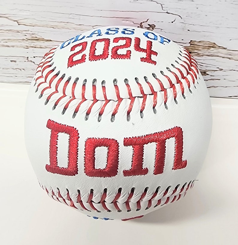 May include: White baseball with red stitching and red embroidered text "Class of 2024" and "Dom"