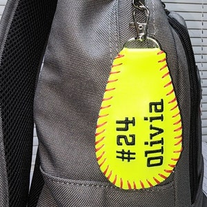 May include: Yellow softball keychain with red stitching and black text that reads "Olivia #24".