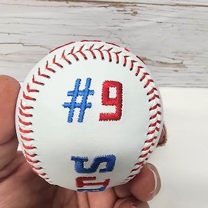 May include: White baseball with red and blue stitching. The number "#9" is embroidered in red and blue on the ball.