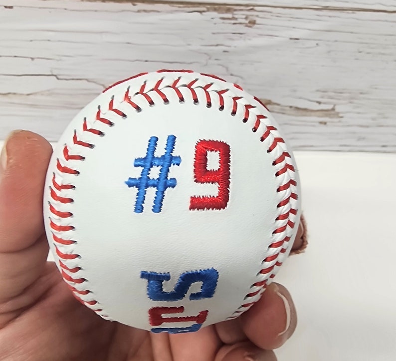 May include: White baseball with red and blue stitching. The number "#9" is embroidered in red and blue on the ball.