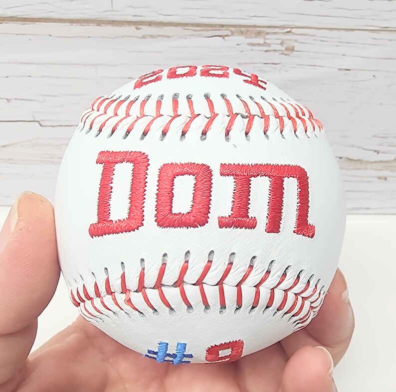May include: White baseball with red stitching and the word "DO IT" embroidered in red. The baseball has a blue number "9" and a blue number "4" on the bottom.