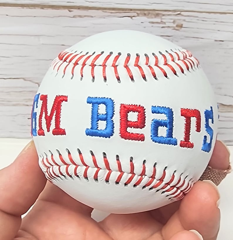 May include: White baseball with red and blue embroidered text that reads "GM Bears".