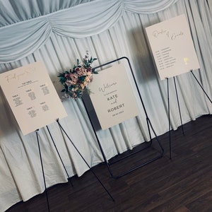 May include: Three white signs with black lettering on black easel stands. The signs are for a wedding and include the order of events, a welcome sign with the names Kate and Robert, and a seating chart titled "Find your Seat".