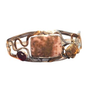 May include: A silver bracelet with a central, speckled, pinkish-brown rectangular stone. The bracelet includes a small, gold-toned turtle and a red gemstone. The design incorporates swirling, vine-like elements, with gold-toned accents.
