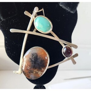 May include: A silver brooch with an abstract design. It features three stones: a turquoise oval, a brown and black patterned oval, and a small, round, dark red stone. The brooch has a geometric, modern aesthetic.