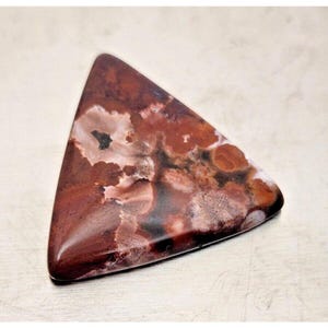 Può includere: Una gemma triangolare lucidata con un mix di colori rosso scuro, marrone e crema. La pietra ha un aspetto marmorizzato con motivi a vortice e una finitura lucida. La forma generale è un triangolo arrotondato.
