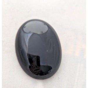 Può includere: Una gemma nera lucida di forma ovale. La pietra ha una superficie liscia e riflettente. La gemma è posta su uno sfondo bianco, evidenziando il suo colore scuro e la sua forma. Misura circa 3 cm di lunghezza. Adatta per gioielli o collezioni.