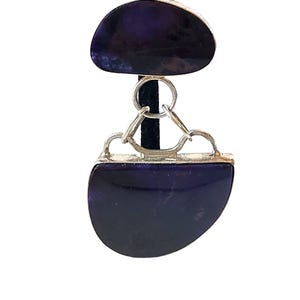 May include: A pendant featuring two polished, deep purple, semi-circular stones connected by a silver-toned metal design. The top stone is smaller than the bottom one. The pendant is displayed against a black background.