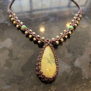 May include: A brown macrame necklace with a teardrop-shaped yellow and green stone pendant. The necklace is accented with small green beads.