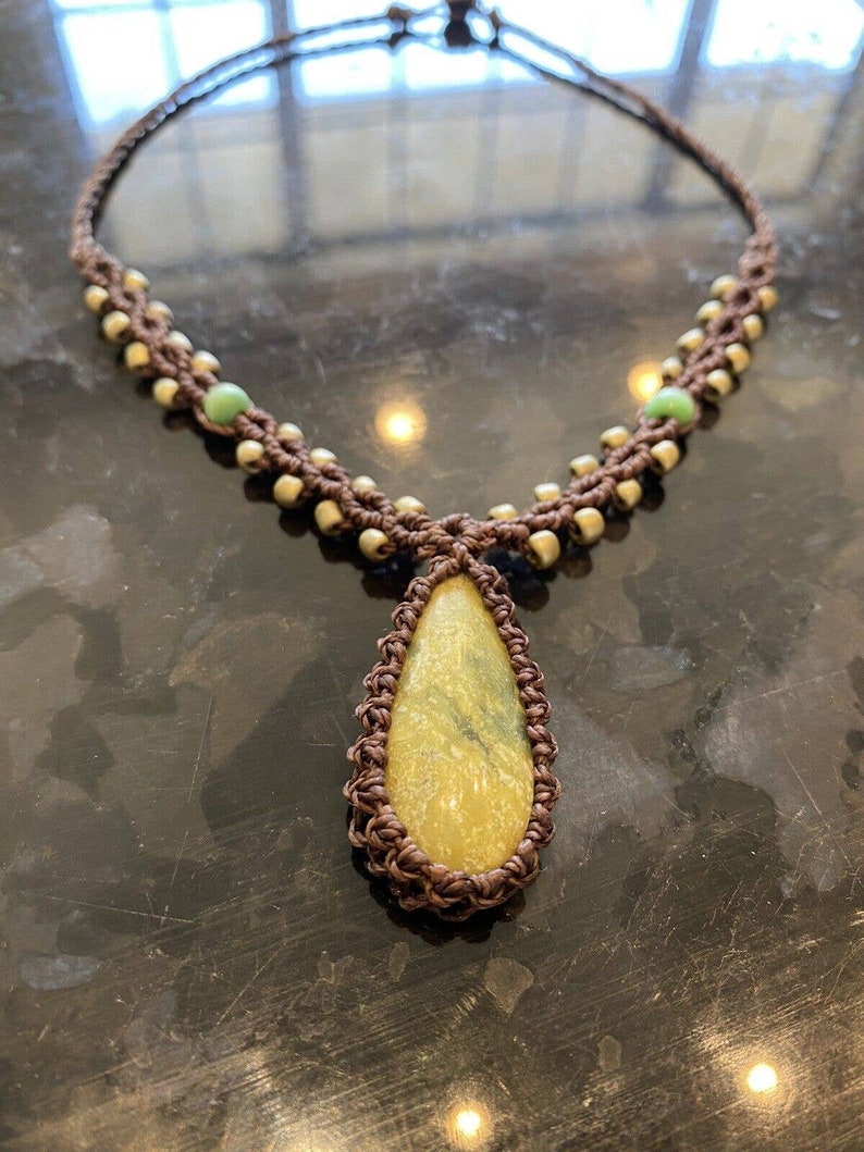 May include: A brown macrame necklace with a teardrop-shaped yellow and green stone pendant. The necklace is accented with small green beads.