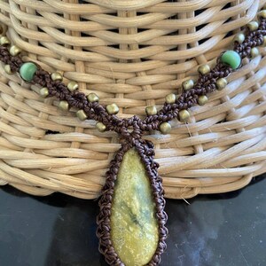 May include: A brown macrame necklace with a green stone pendant. The necklace has gold beads and green glass beads.