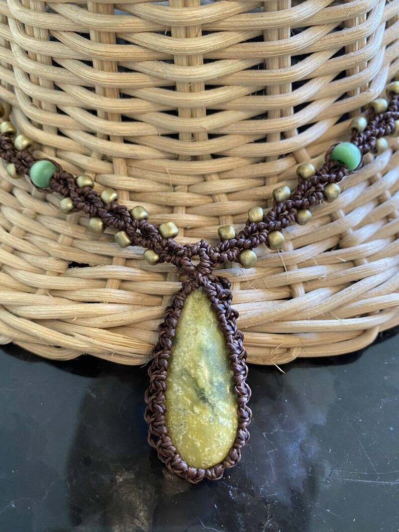 May include: A brown macrame necklace with a green stone pendant. The necklace has gold beads and green glass beads.