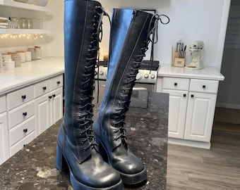 May include: A pair of black knee-high boots featuring a full-length lace-up front with black laces, a side zipper closure, a rounded toe, and a chunky block heel with a platform sole. The boots are constructed from a smooth black material.