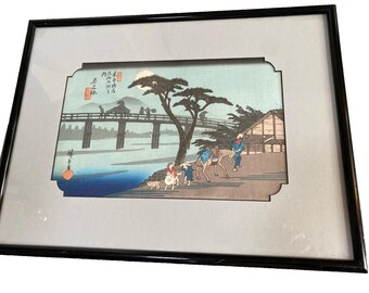 May include: Framed Japanese woodblock print depicting a landscape with a bridge, figures, and a tree. The artwork features a muted color palette with a black frame and a white mat. The print includes Japanese text.