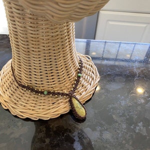 May include: A brown and green beaded necklace with a large green stone pendant. The necklace is displayed on a wicker mannequin.