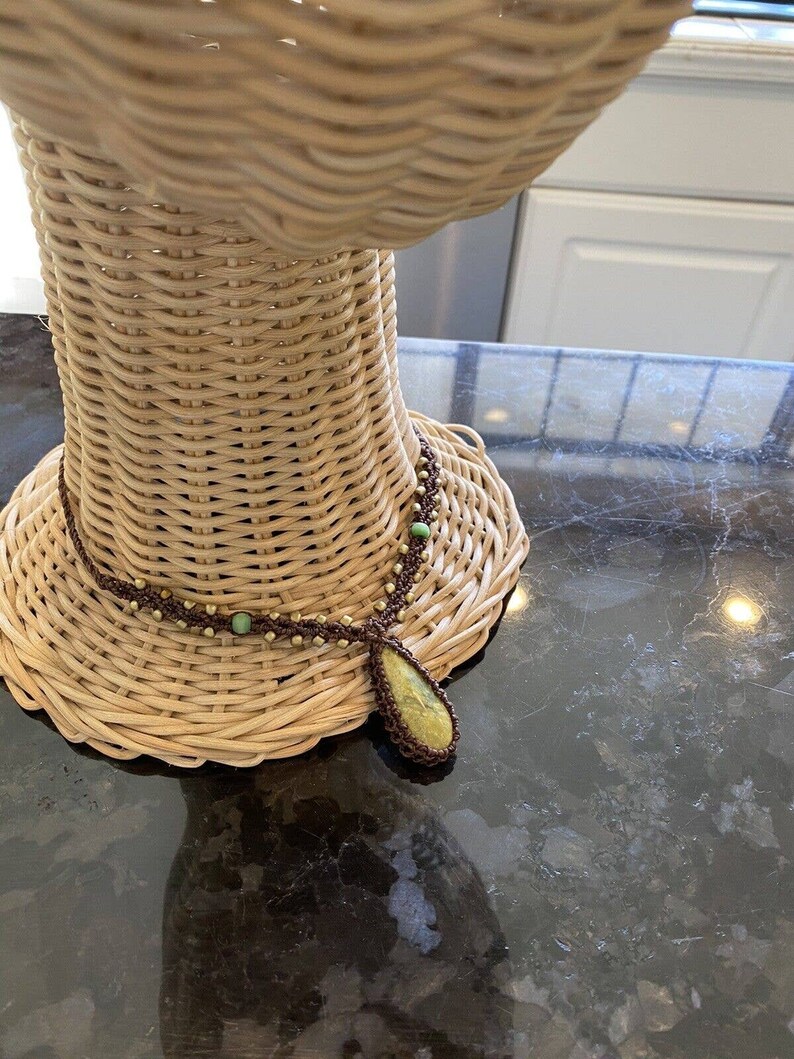 May include: A brown and green beaded necklace with a large green stone pendant. The necklace is displayed on a wicker mannequin.