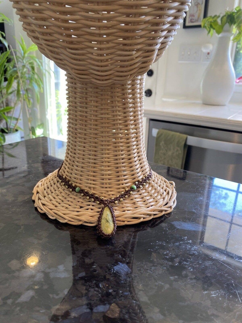 May include: A brown and green beaded necklace with a large teardrop-shaped yellow stone pendant. The necklace is displayed on a wicker mannequin head.