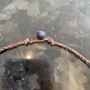 May include: A close-up of a brown rope necklace with a dark blue bead. The necklace is tied in a knot and the bead is centered on the knot.
