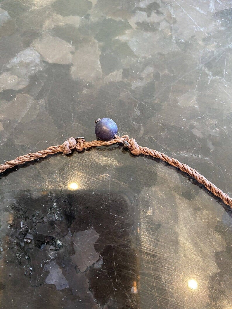 May include: A close-up of a brown rope necklace with a dark blue bead. The necklace is tied in a knot and the bead is centered on the knot.