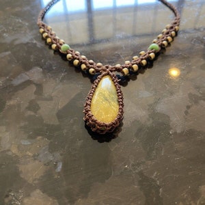May include: A brown macrame necklace with a yellow and green gemstone pendant. The necklace is made with small brown and white beads.