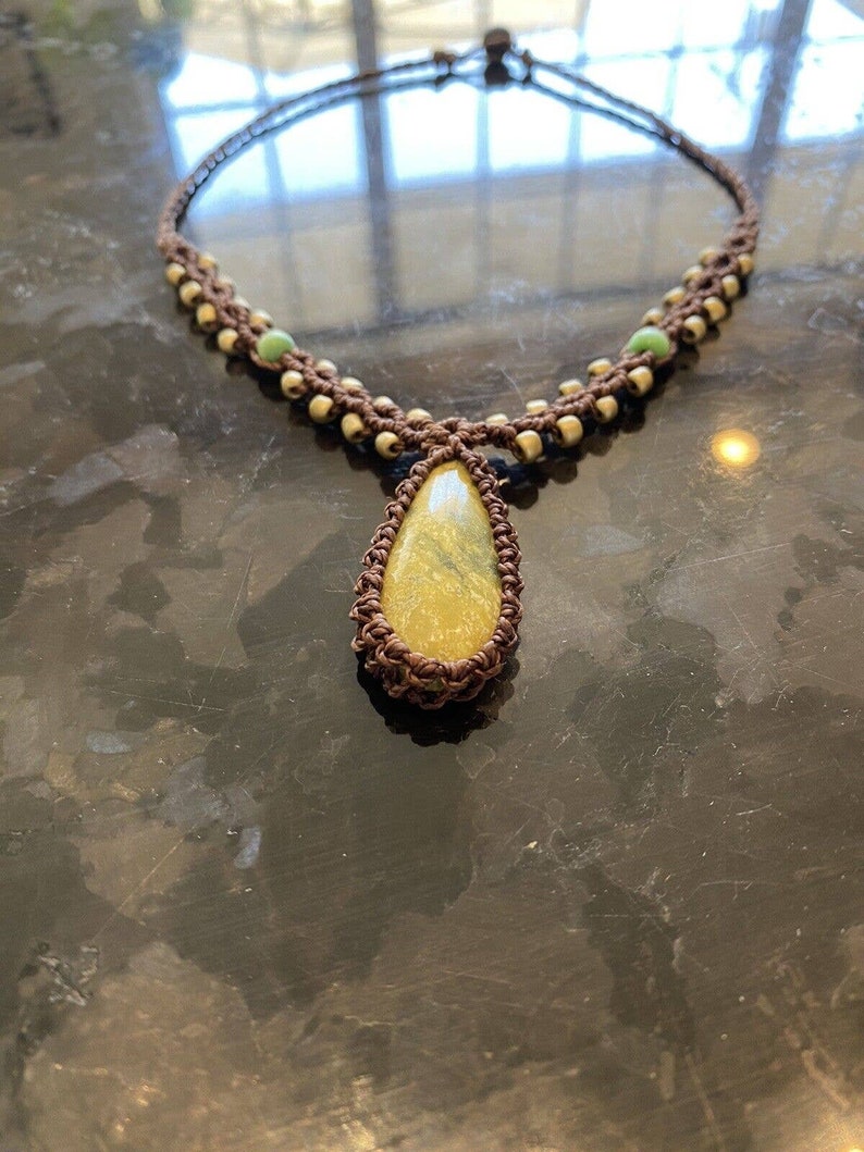 May include: A brown macrame necklace with a yellow and green gemstone pendant. The necklace is made with small brown and white beads.