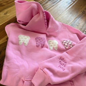 May include: Pink hooded sweatshirt with three appliqued teeth and the embroidered text "future tooth fairy" on the sleeve. The teeth have different patterns and colors. The sweatshirt has a front pocket and ribbed cuffs.