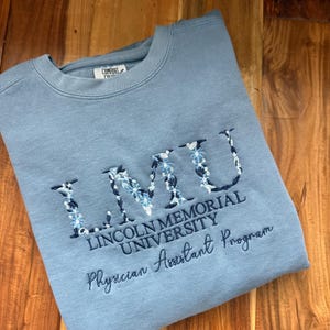 May include: A light blue sweatshirt with embroidered text. The text reads "LMU LINCOLN MEMORIAL UNIVERSITY Physician Assistant Program". The letters of "LMU" are filled with a floral design in shades of blue and white.