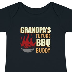 May include: A dark blue baby onesie with the text "Grandpa's Future BBQ Buddy" in yellow and orange. A red flame with a grill and a piece of meat is above the text.