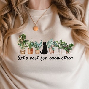 May include: A beige sweatshirt with a black cat sitting among potted plants. The text "Let's root for each other" is printed below the plants.