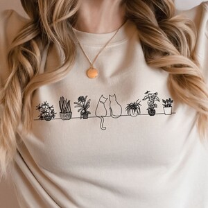 May include: A cream-colored sweatshirt with a black line drawing of two cats sitting amongst potted plants. The text "Sand" is visible in the bottom right corner of the image.