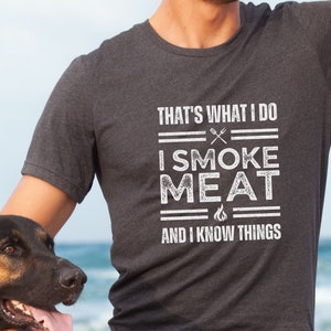 May include: A man wearing a dark grey t-shirt with the text "That's what I do I smoke meat and I know things". He is standing next to a German Shepherd dog.