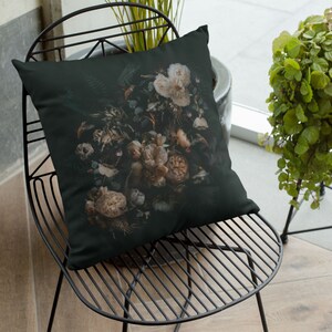 May include: A decorative throw pillow featuring a dark background with a detailed floral arrangement. The flowers are primarily cream and beige, with hints of green foliage. The pillow is square and rests on a black metal chair.