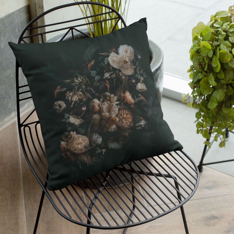 May include: A decorative throw pillow featuring a dark background with a detailed floral arrangement. The flowers are primarily cream and beige, with hints of green foliage. The pillow is square and rests on a black metal chair.