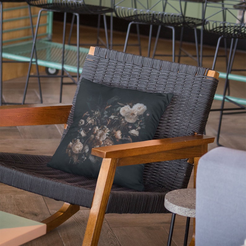 May include: A decorative throw pillow with a dark background and a floral design featuring white and beige flowers. The pillow is placed on a gray woven chair with wooden armrests, creating a contrast of textures and colors.