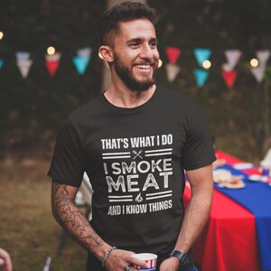 May include: A black t-shirt with white text that reads "That's what I do I smoke meat and I know things". The text is stylized with lines and a grill icon.