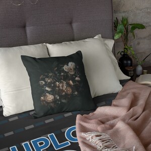 May include: A decorative throw pillow with a dark background featuring a floral still life in shades of cream, brown, and green. The pillow is square and rests on a bed with white pillows and a patterned bedspread. A pink blanket is draped over the bed.