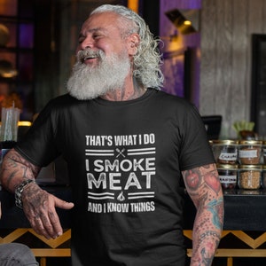 May include: A black t-shirt with a white graphic that says "That's what I do I smoke meat and I know things". The graphic includes a grill, a knife, and a flame.