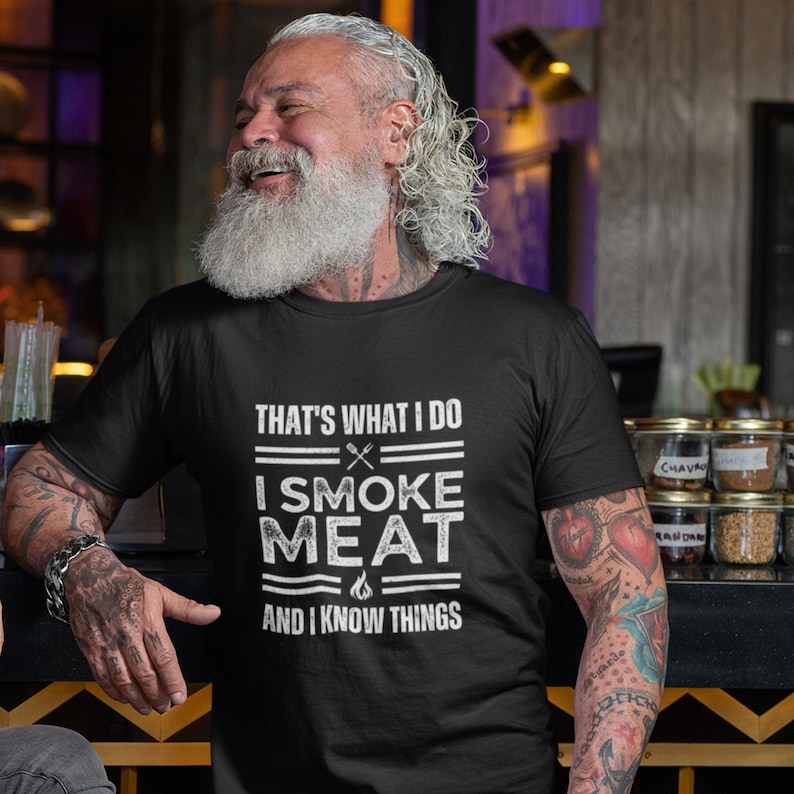 May include: A black t-shirt with a white graphic that says "That's what I do I smoke meat and I know things". The graphic includes a grill, a knife, and a flame.