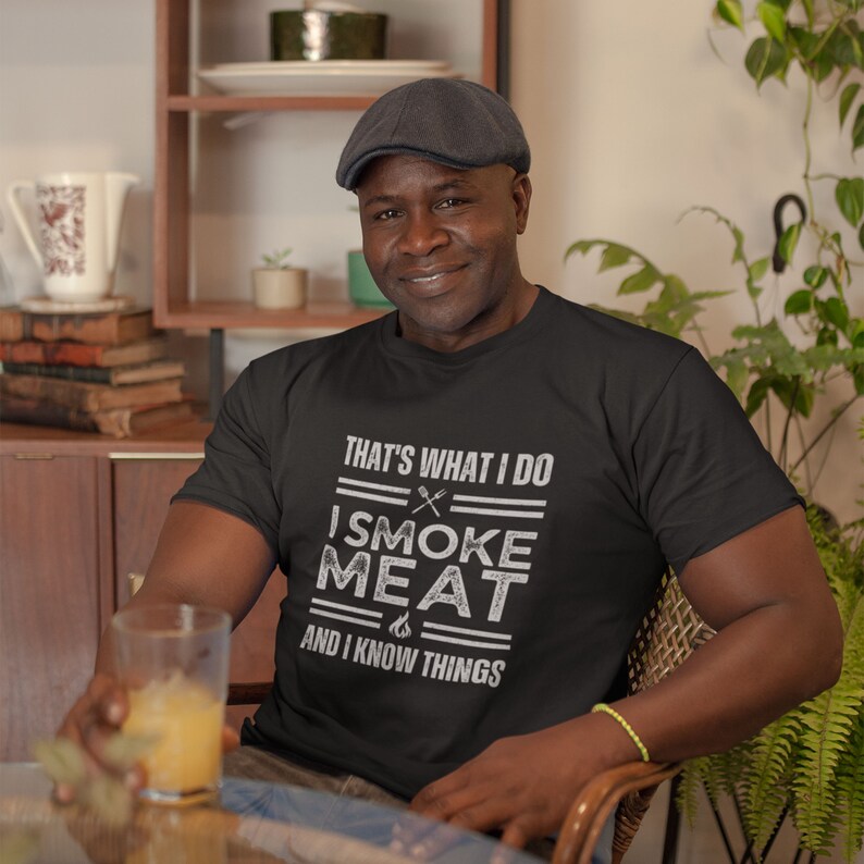 May include: A man wearing a black t-shirt with the text "That's what I do I smoke meat and I know things".